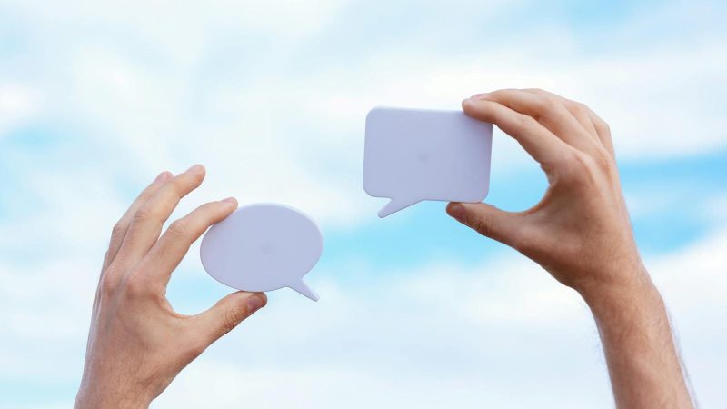 Close-up of hands holding speech bubbles, symbolizing communication and dialogue against a blue sky.