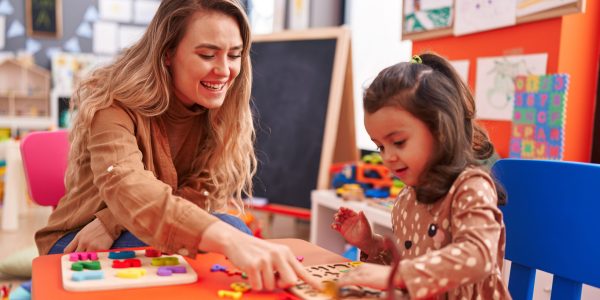 Teacher and toddler playing with maths puzzle game sitting on table at kindergarten
